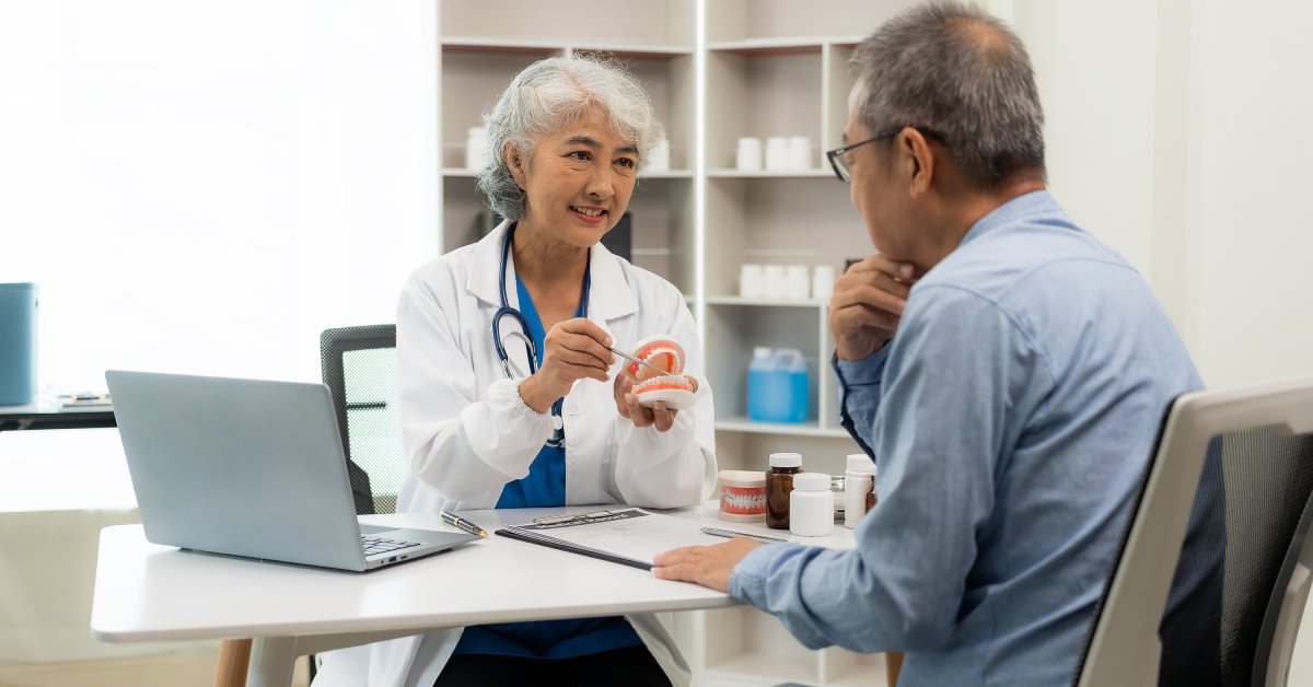 An older female dentist sitting at a desk shows an older man a dental model with a device in a medical office.