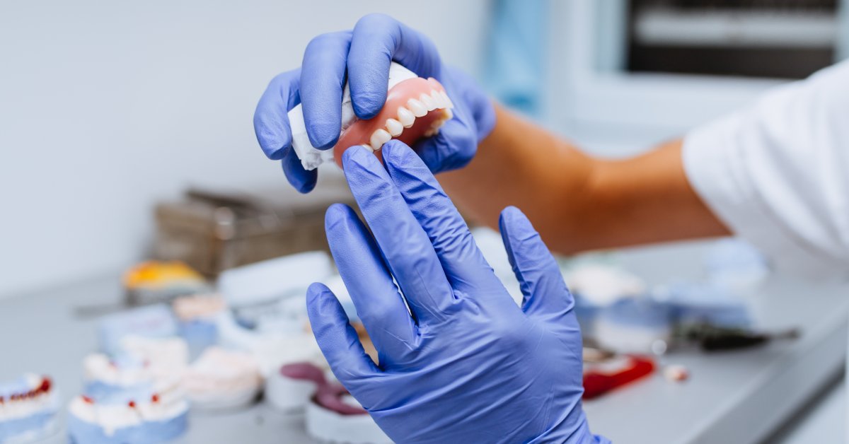 A person's hands in blue gloves are displaying a dental model with medical equipment sitting on a table in the background.