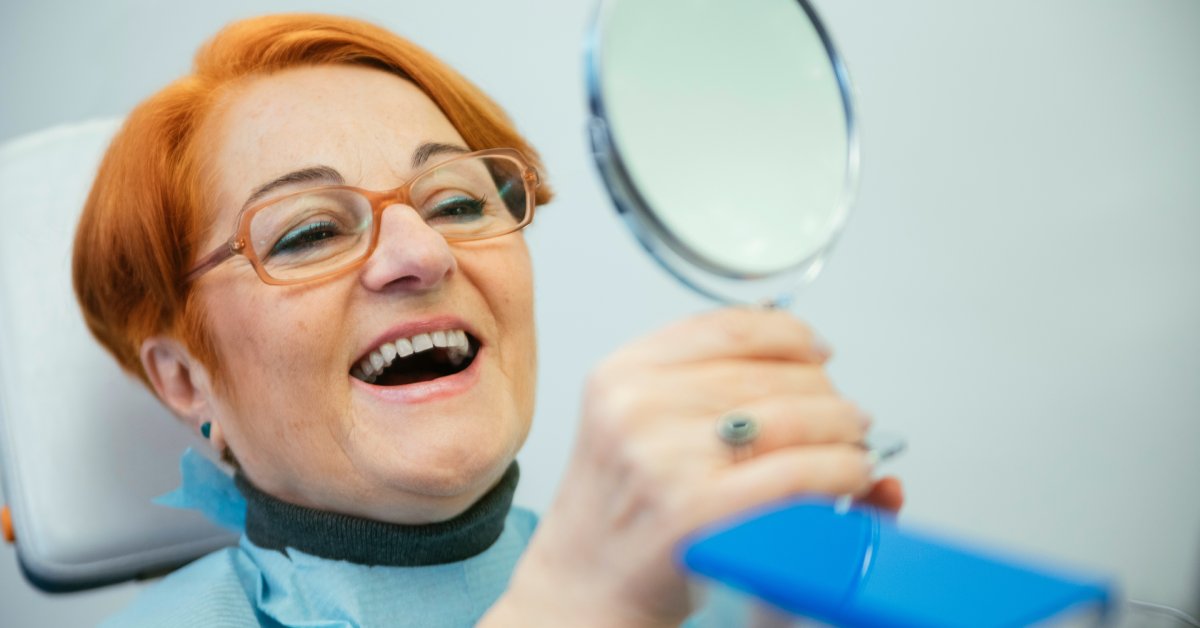 An older woman wearing glasses and a dental bib is sitting in a medical chair and looking at her smile in the mirror.