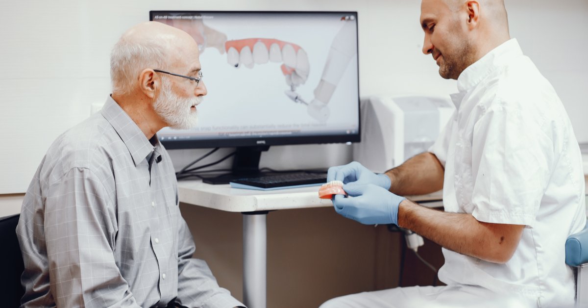 An older male is sitting in front of a screen displaying a dental model and looking at dentures with a dentist.