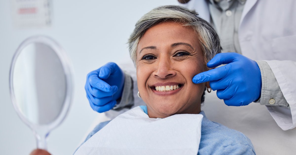 An older female patient holds a mirror and smiles with her teeth while sitting in front of a dentist with gloved hands.