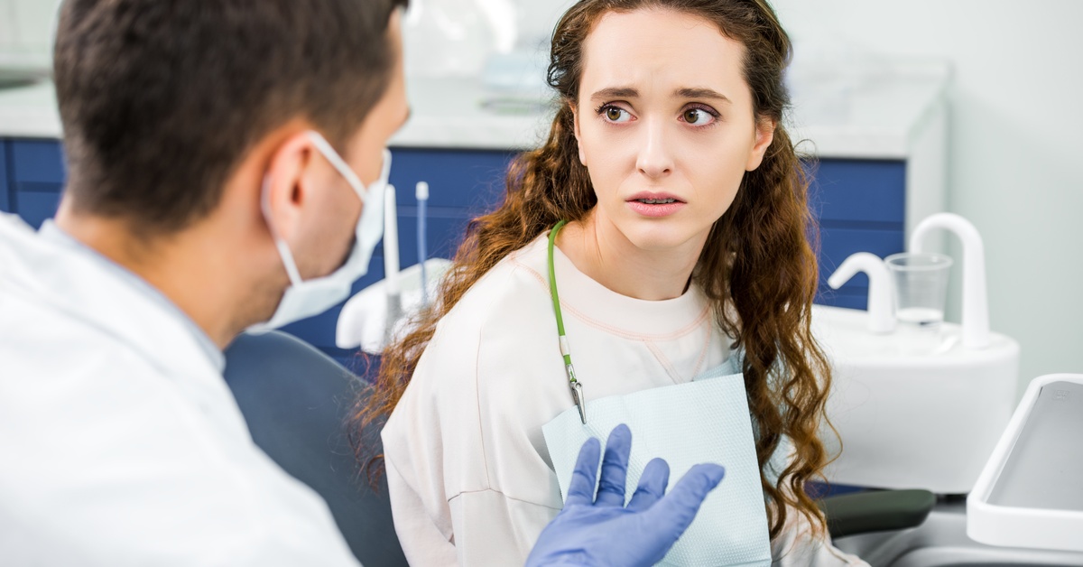 A woman with a worried expression sits in a dentist's office, talking to a masked dentist wearing gloves.