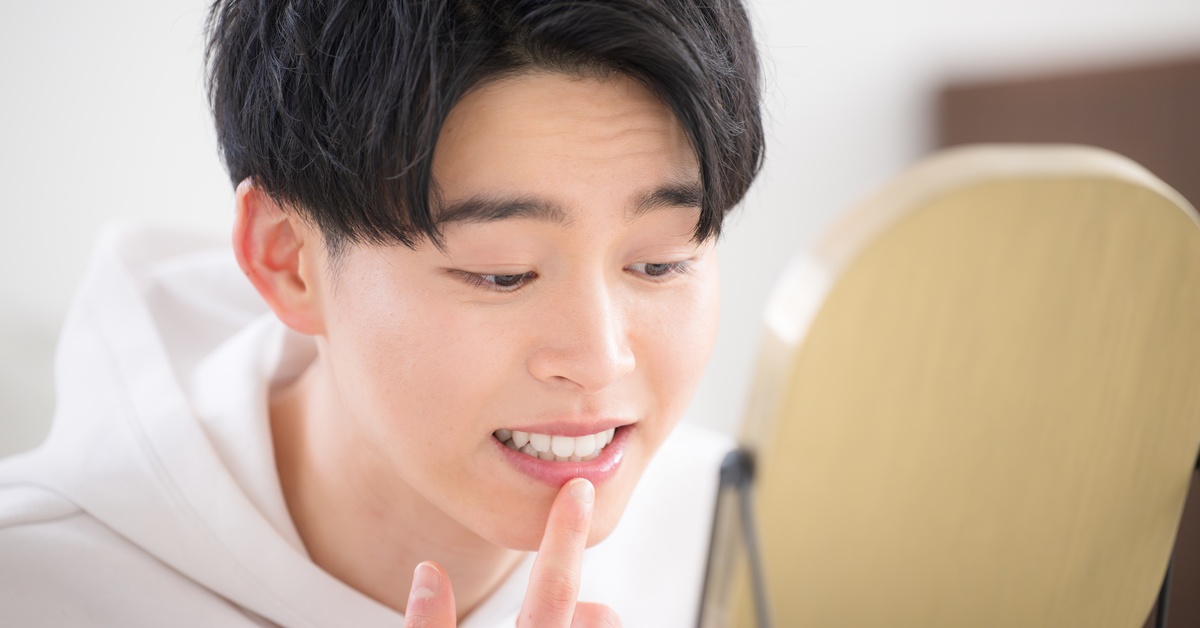 A man in a white hoodie is looking at himself in a small mirror while smiling and touching his lower lip.