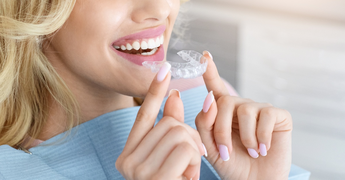 A woman with an open-mouth smile and wearing a dental bib holds a set of clear dental aligners between her fingers.