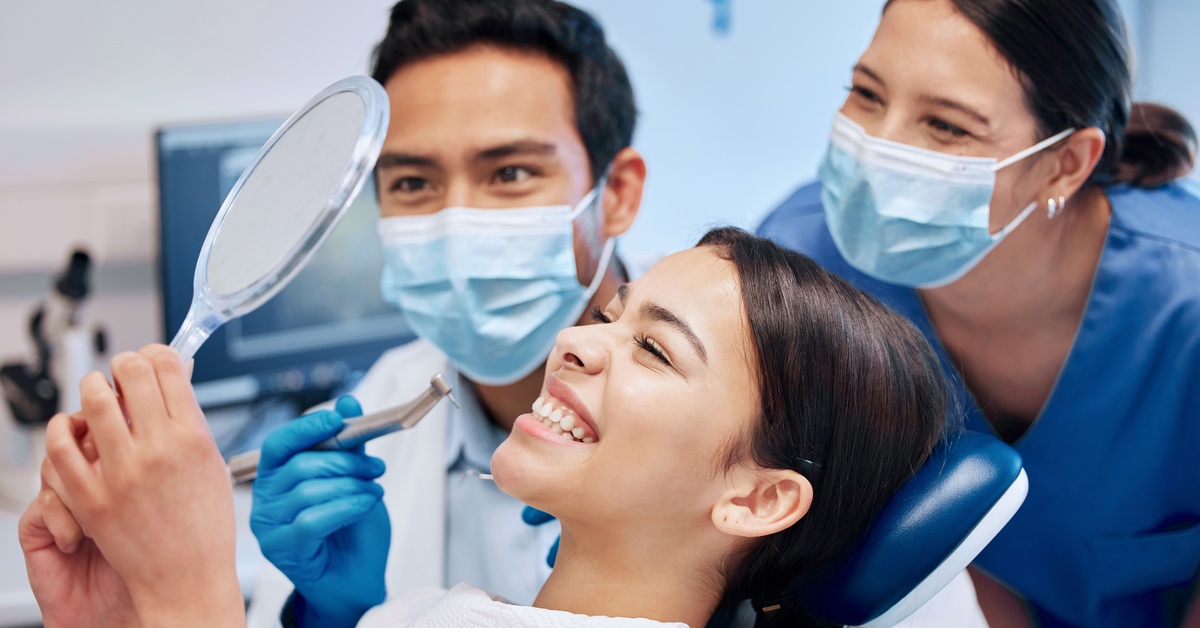 A woman wearing a dental bib smiles with her teeth while looking in the mirror, with two dental professionals observing.