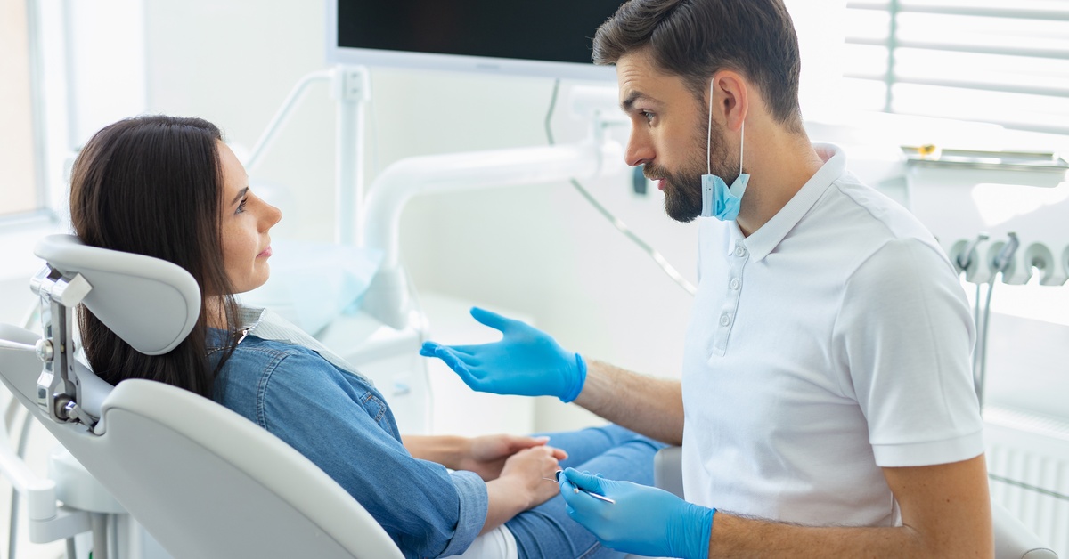 A male dentist with gloves and a mask is talking with a serious expression to a female sitting in a dental patient chair.