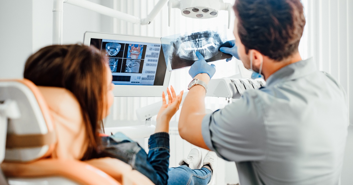 A male dentist is showing imaging from scans of teeth, pointing to it alongside a female sitting in the patient chair.