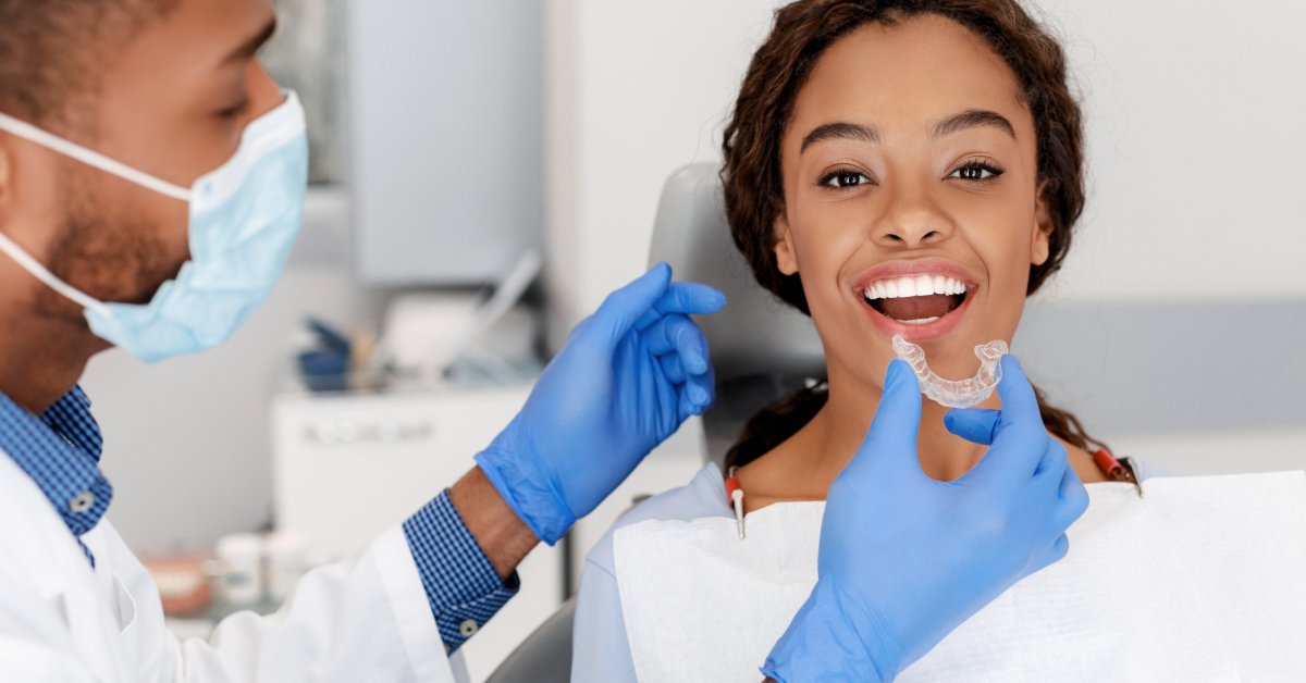A woman is sitting in a dental office in the patient chair with a masked dentist with gloves holding an aligner to her mouth.