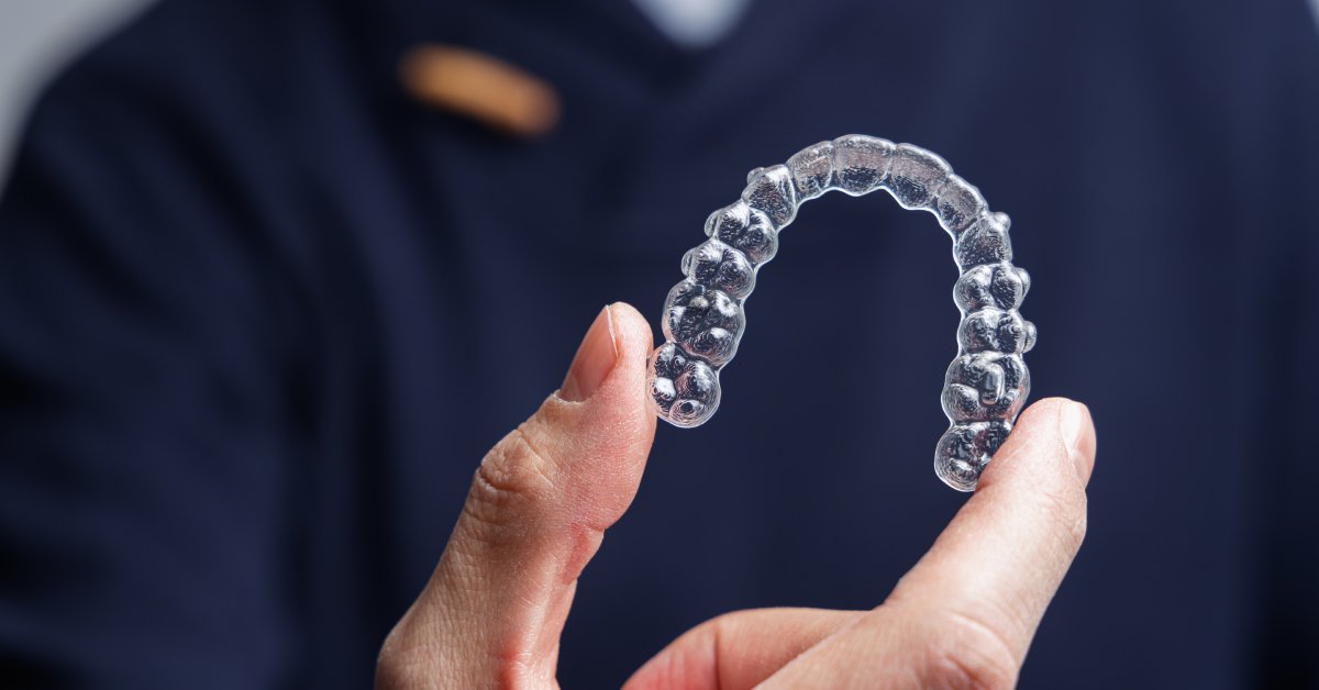 A man in dark blue scrubs with his face out of frame is holding up a set of transparent teeth aligners in his hand.