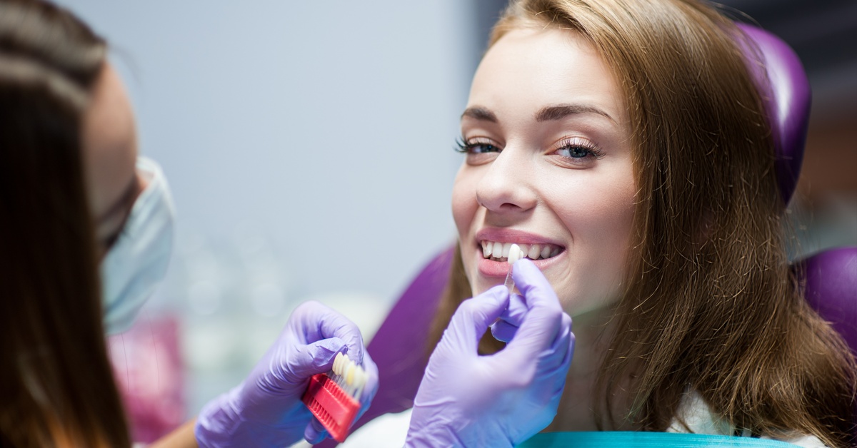 A patient is sitting in a patient chair, smiling as a gloved dentist matches enamel color options to the patient's teeth.