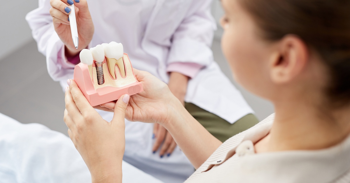 A patient holds a model of a dental implant while a dentist uses a pointer to point alongside the dental model.