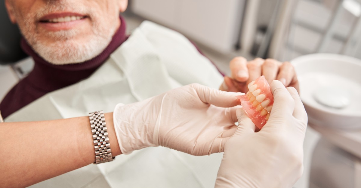 A person wearing medical gloves shows a male patient in a dental bib a pair of dentures. They're in a medical office.