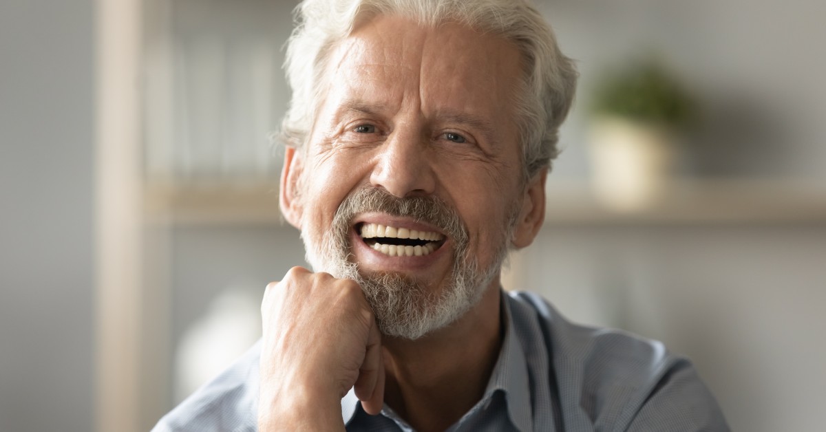 An older man is smiling with his mouth open, displaying his teeth, and resting his hand under his chin in a pose.