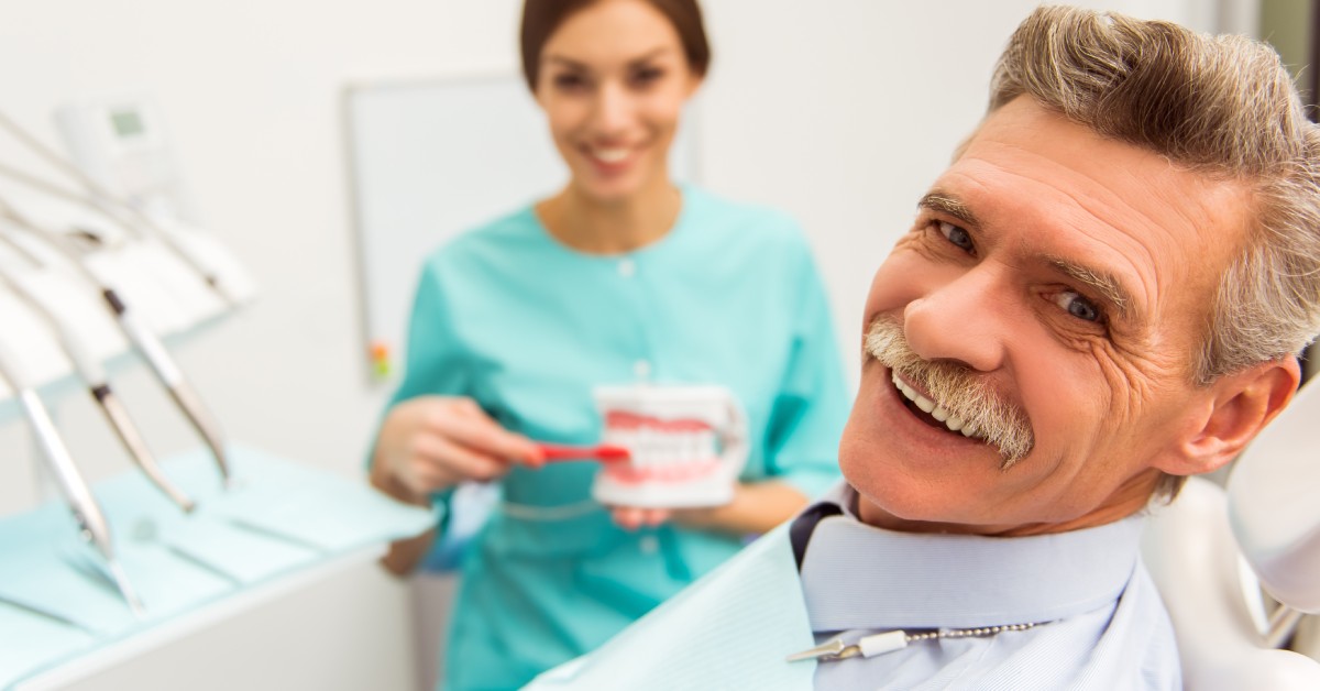 An older man in a dental office is smiling, while a dentist in scrubs holds a dental model in the near distance.