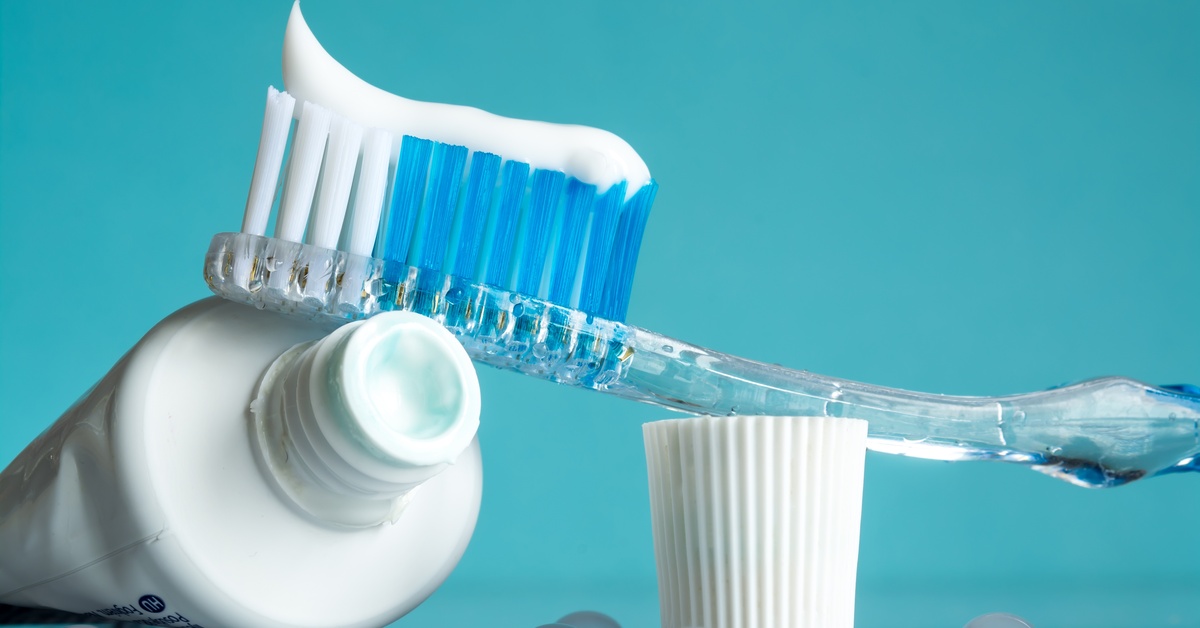 A toothbrush with a line of toothpaste on it is sitting on top of an open tube of toothpaste, with a blue background.