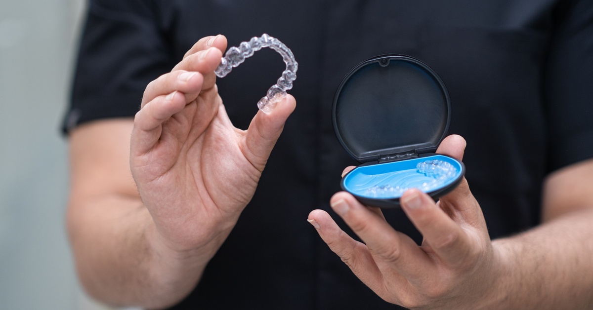 A close-up of a man in dark scrubs holds a clear tooth aligner in one hand and a tooth aligner case in the other.