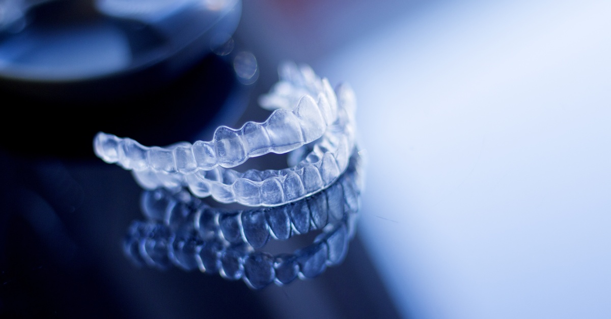 A close-up of clear teeth aligners stacked on top of one another on a shiny, reflective, dark blue surface.