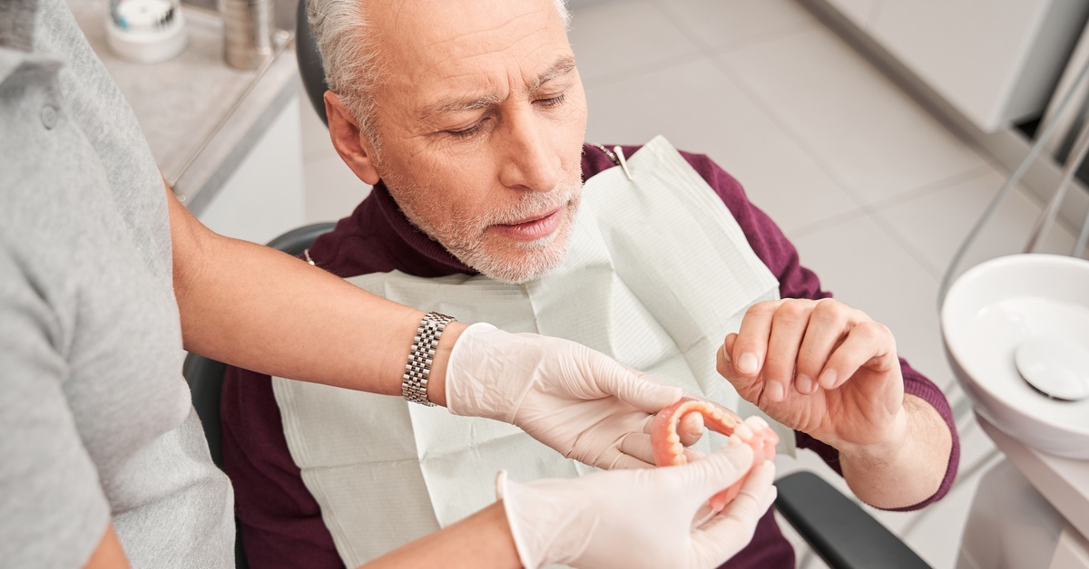 A man wearing a dental bib sits in a dentist's chair. A person wearing latex gloves is showing him a set of dentures.