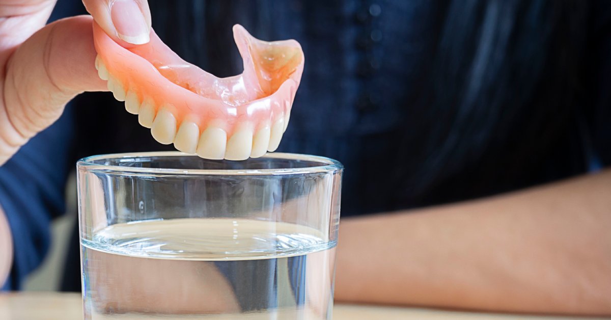 A close-up of a person's hand holding a top denture piece over a small glass of water for cleaning and soaking.