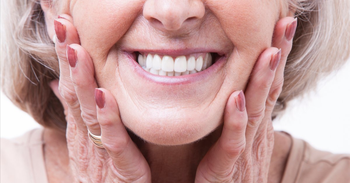 A close-up of a person smiling wide with their straight, white teeth showing. Their hands are placed on either side of their face.