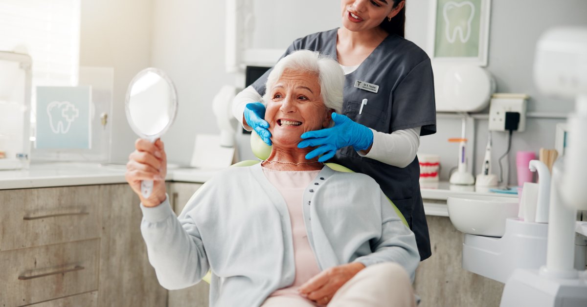 An older woman is sitting in a dentist's chair and smiling into a handheld mirror. A dentist stands behind her wearing scrubs.