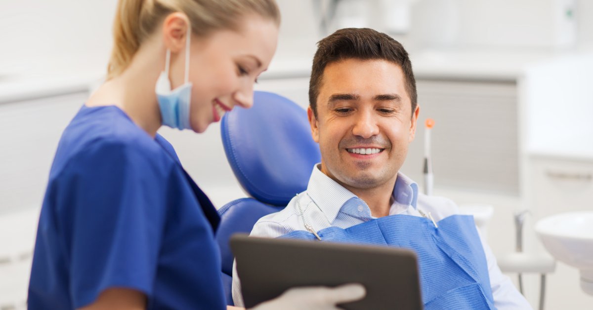 A man sits in a dental exam chair as a dentist shows him something on a digital tablet. They're both smiling.