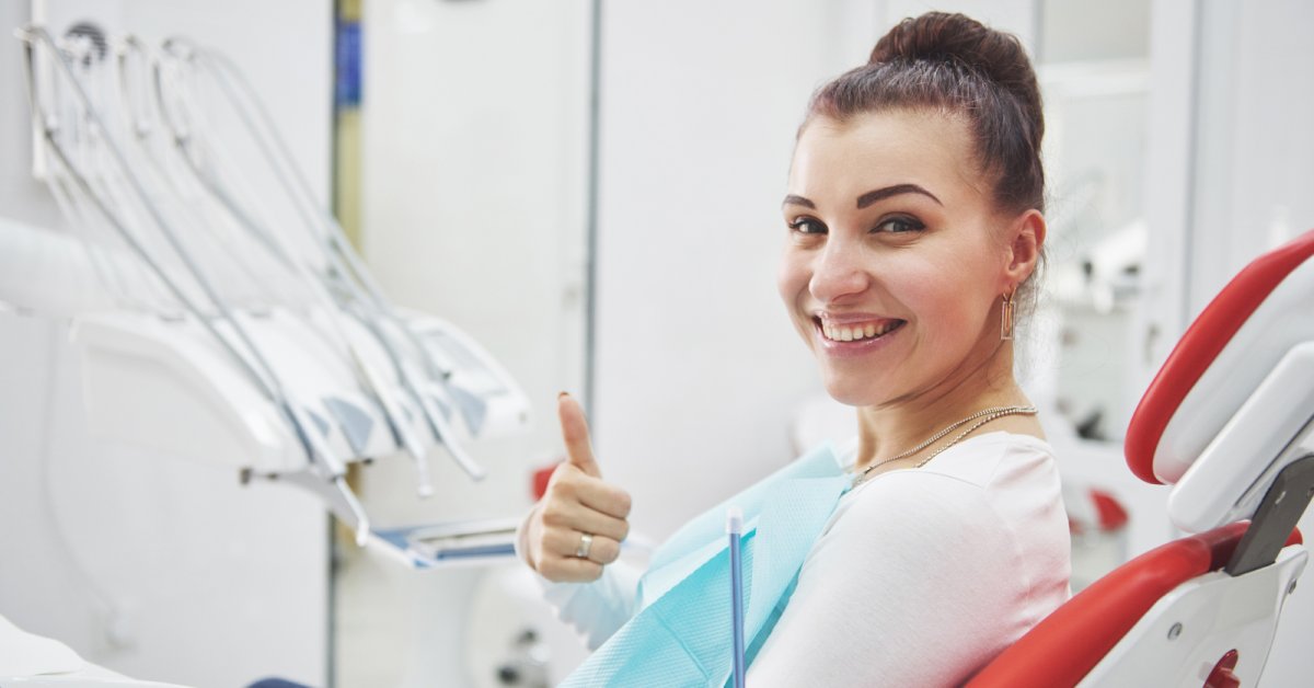 A woman sits in a dental exam chair and smiles widely. She's wearing a dental bib and giving a thumbs up.
