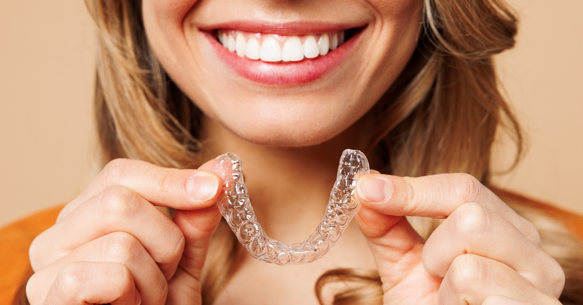 A close up of a person smiling widely, showing their teeth. They're holding a clear aligner in front of them.