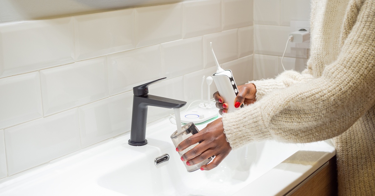A person fills the reservoir of a water flosser at a bathroom sink. They're holding the flosser in their other hand.