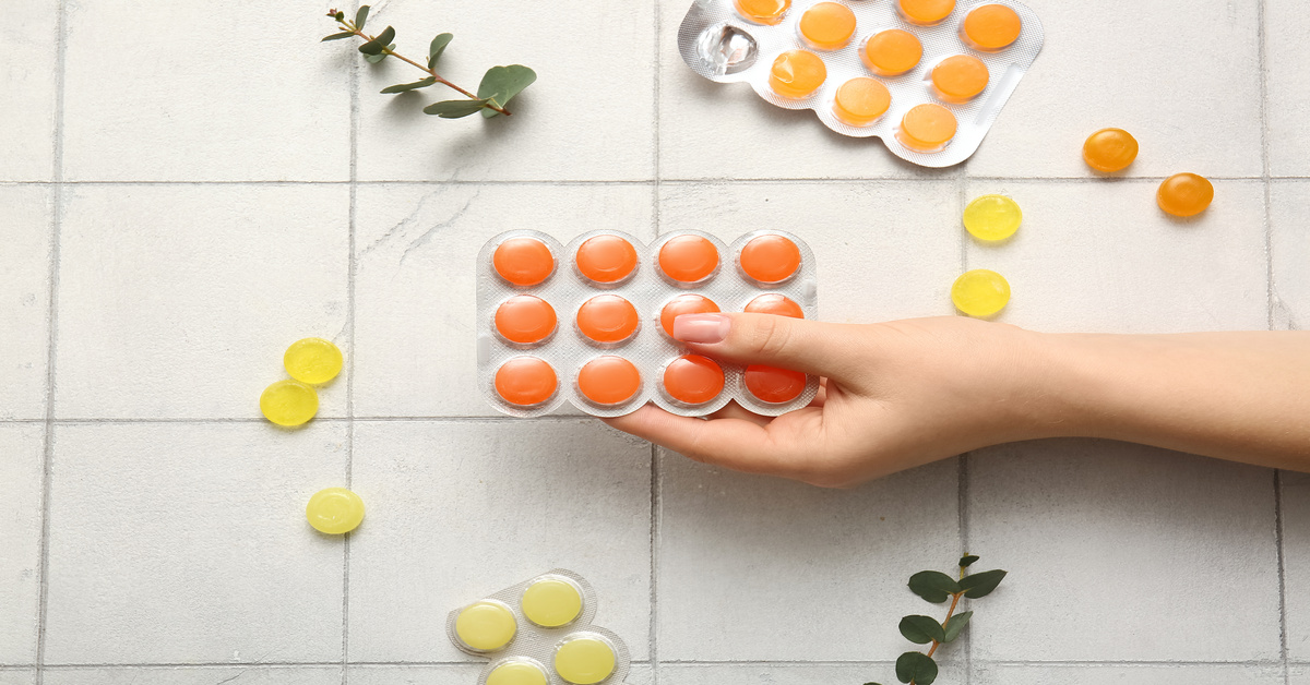 A hand holding a package of pink cough drops against white tile. Eucalyptus leaves and cough drops lie on the tile.