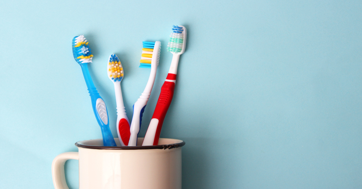 Four toothbrushes in different colors and sizes sit in a white ceramic mug against a light blue wall.