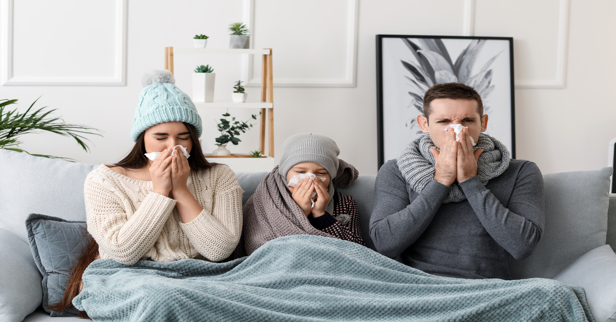 A family of three sits on a couch wrapped in scarves and blankets. They're all holding tissues to their noses.