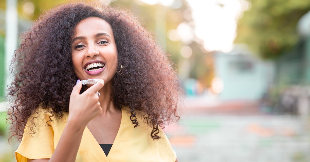 A woman smiles confidently as she holds a clear aligner up to her mouth. She's standing outside on a sunny day.