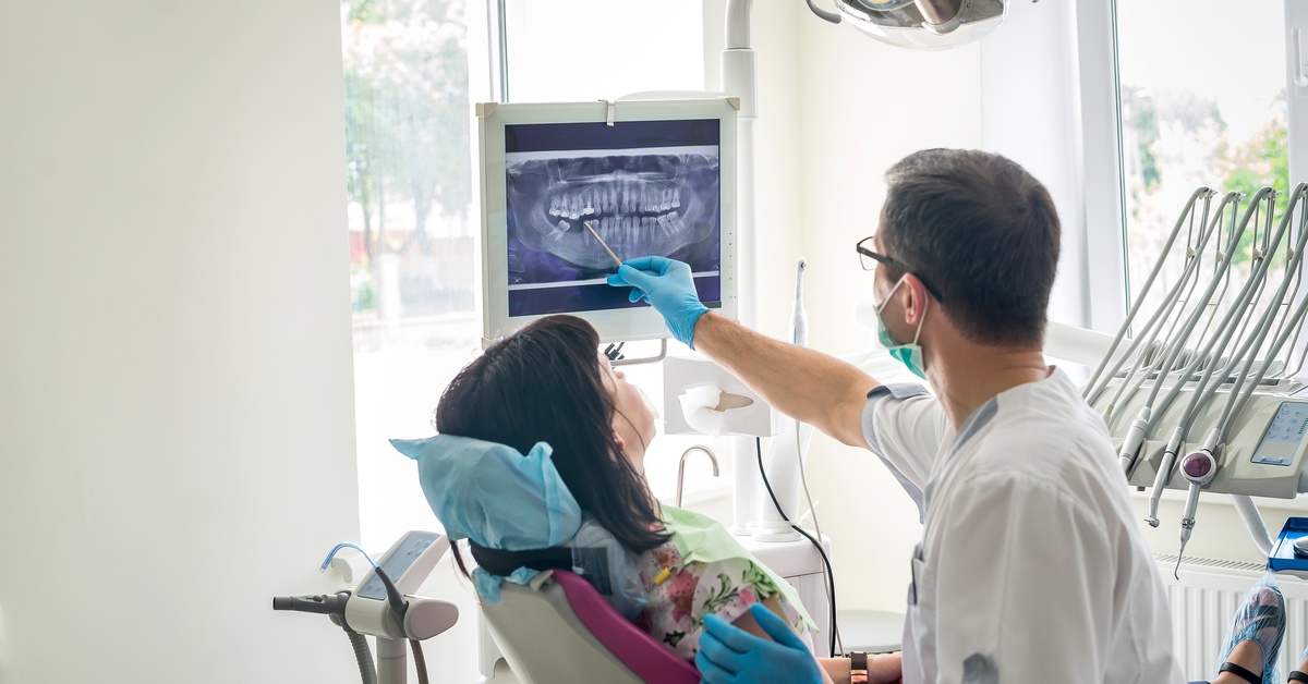 A dentist shows a patient in an exam chair an X-ray of their teeth on a screen. The dentist is pointing at the X-ray with a utensil.
