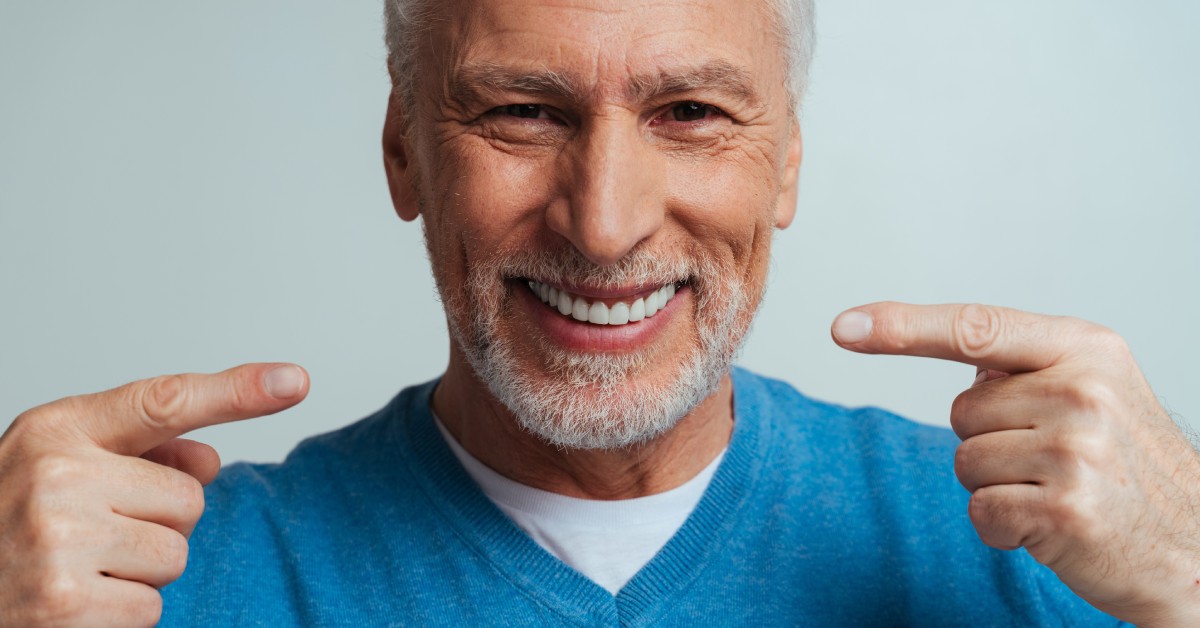 An older man wearing a blue sweater smiles widely, showing his teeth. He is confidently pointing to his mouth.