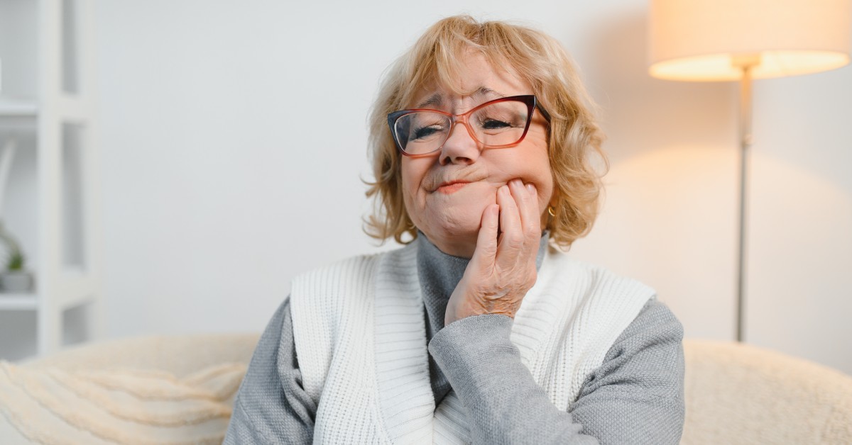 An older woman wearing a sweater vest and glasses holds the side of her face and screws her mouth to the side in discomfort.