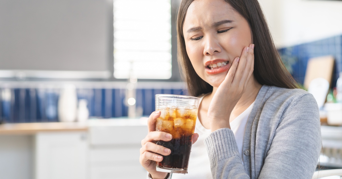 An Asian woman touches the side of her face as she grimaces in pain. She is holding a cold beverage in her hand.