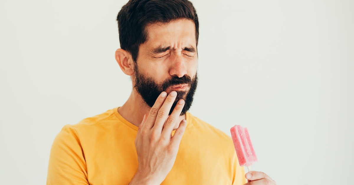 A man in a bright yellow shirt is holding a popsicle and touching his mouth with his other hand as he grimaces in pain.