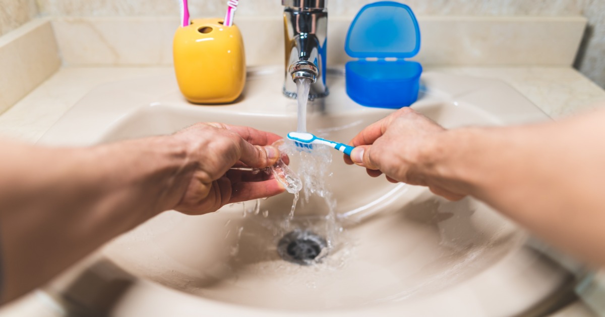 A person uses a toothbrush to scrub a clear aligner under a faucet. A toothbrush holder and aligner case sit on the sink.