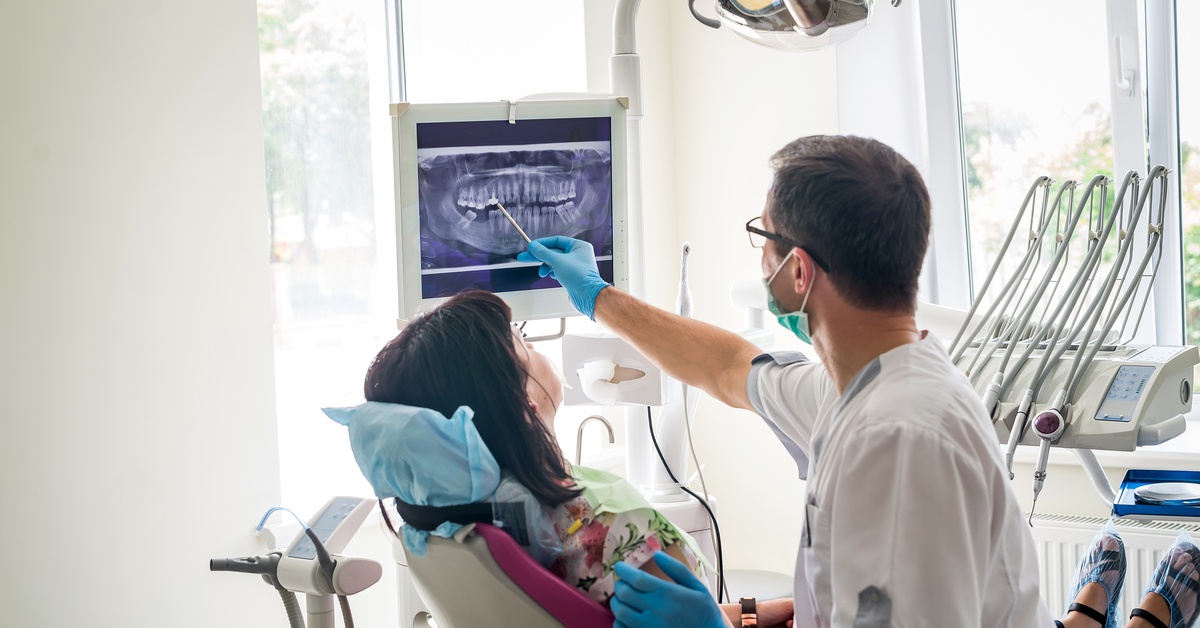 A male dentist in an exam room points to an X-ray on a monitor while a female seated patient listens.