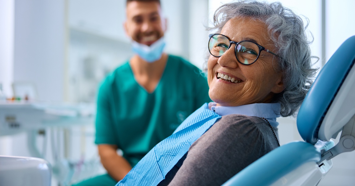 A smiling older woman sits in a dental chair wearing a bib while a dentist in scrubs sits behind her in an exam room.