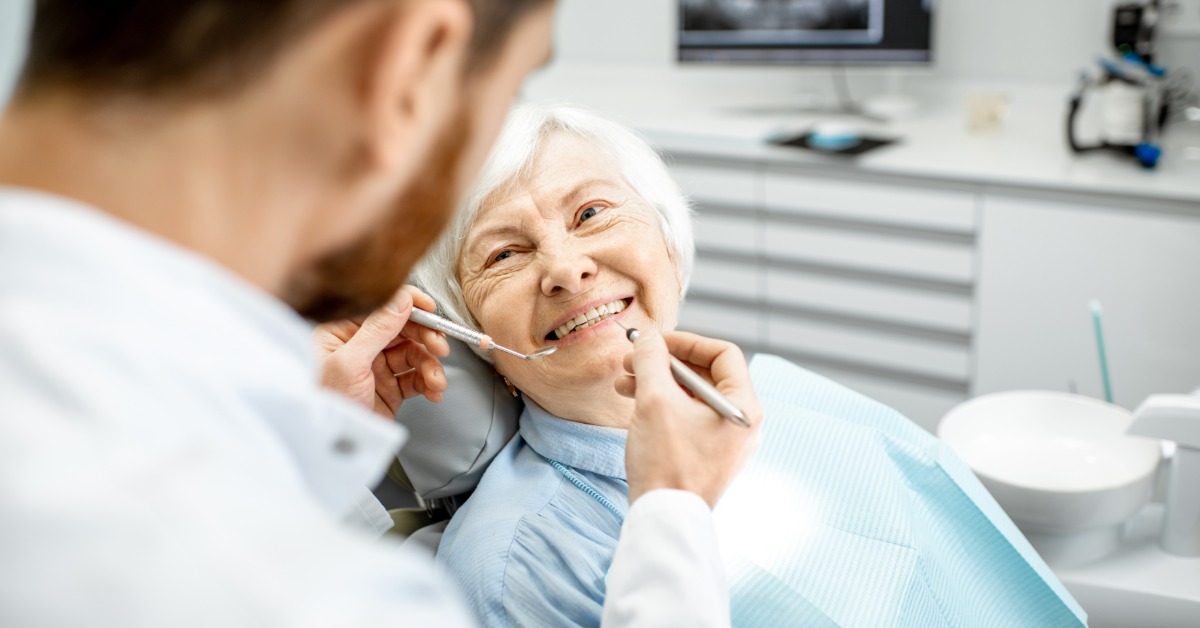 A male dentist uses two dental tools to examine a smiling elderly woman sitting in a chair in a modern clinic.