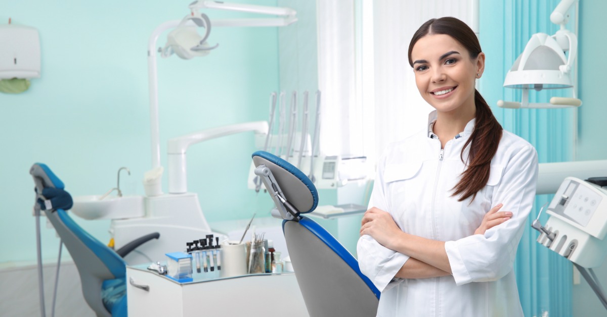 A smiling female dentist stands in a modern dental office with chairs, tools, and equipment visible in the background.
