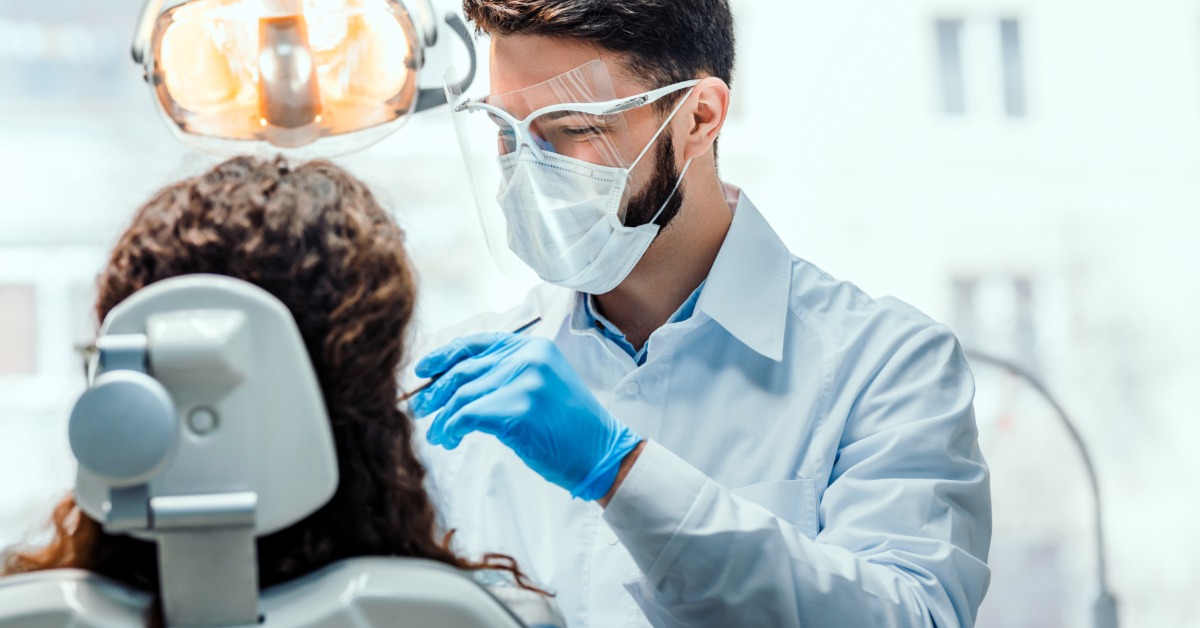 A dentist wearing a mask and face shield examines a patient in a dental chair under bright overhead light.