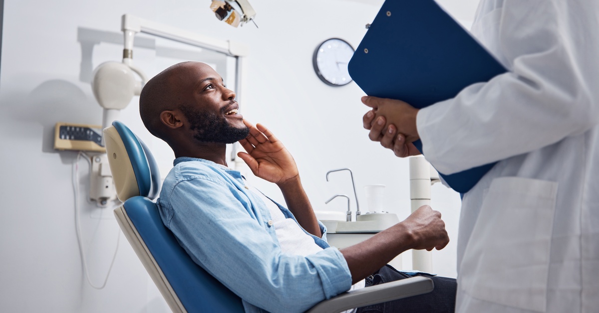 A man sits in a dentist's exam chair and smiles up at the dentist, who is standing in front of him and holding a clipboard.