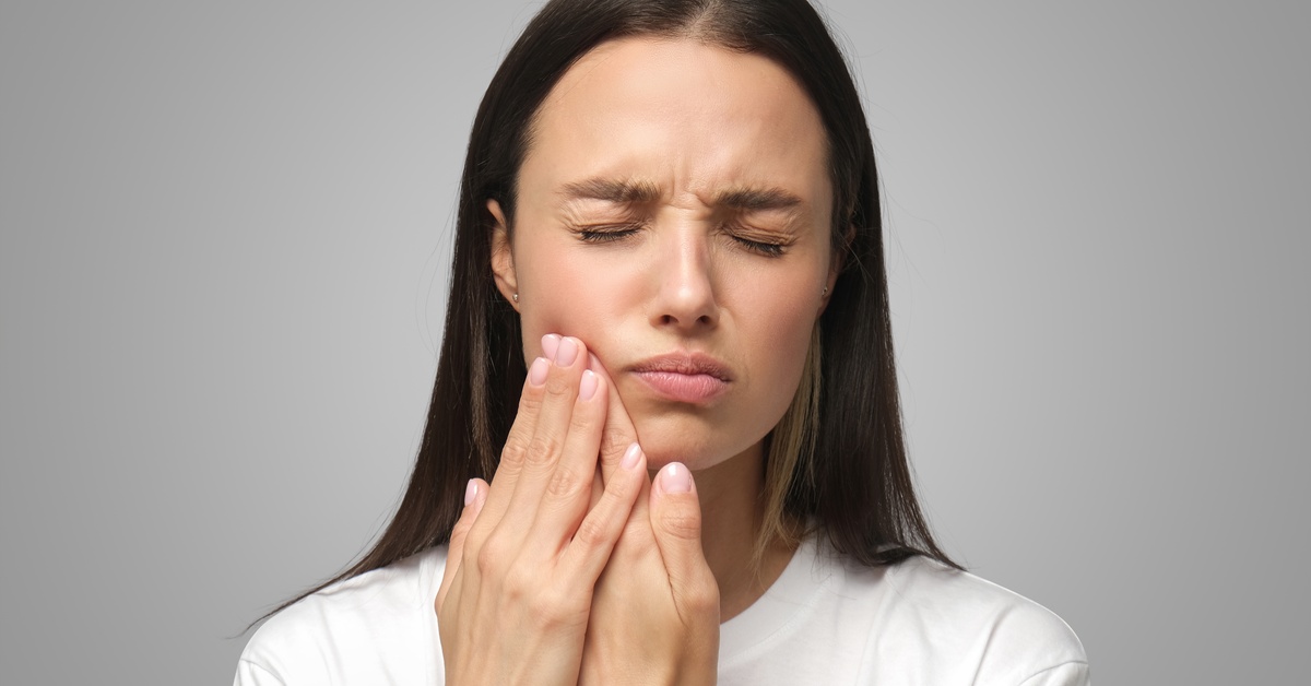 A woman with brown hair grimaces in pain as she holds the side of her face with both hands against a gray background.