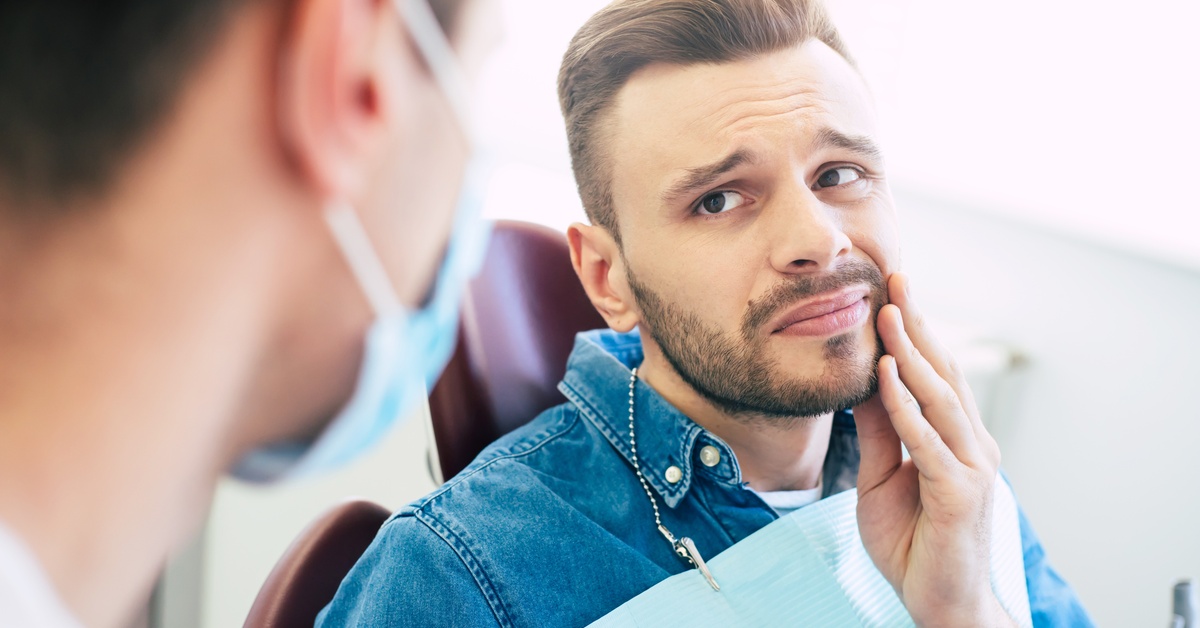 A man in a dental chair holding his cheek in discomfort while speaking to a dentist wearing a mask during an exam.