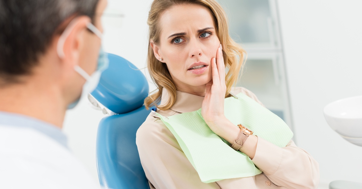 A woman in a dental chair holding her cheek in pain while looking at a dentist wearing a mask during an exam.