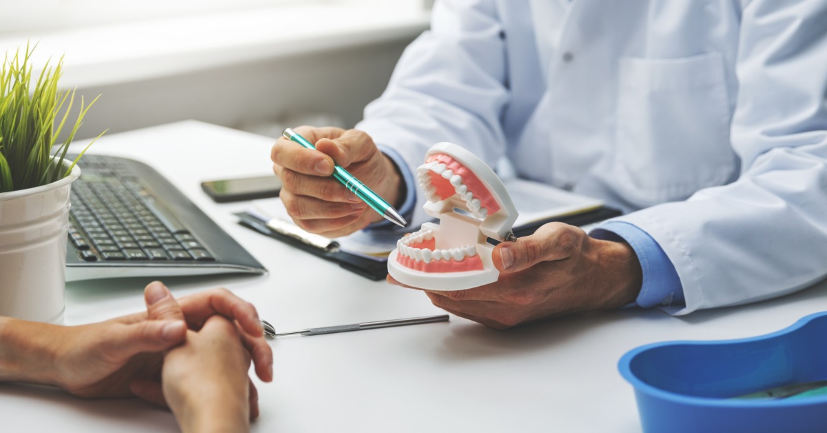 A dentist holding a dental model while explaining treatment options to a patient seated across the desk.