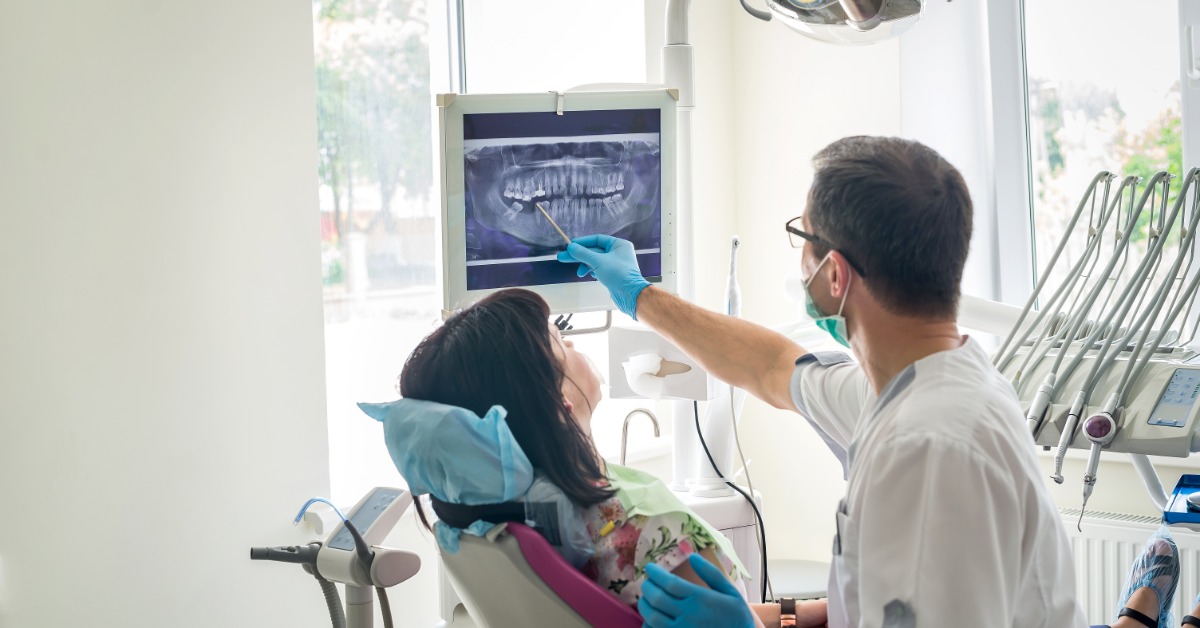 A dentist points to a dental X-ray on a monitor while discussing treatment with a patient in a dental chair.